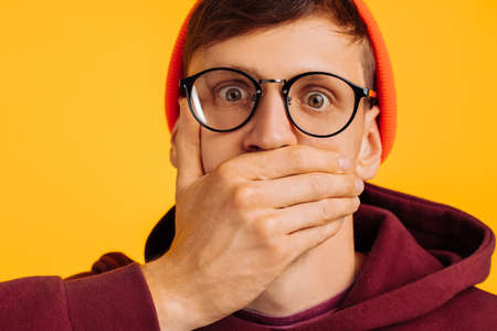man covered his mouth with his hand, a frightened man, in an orange hat and red sweatshirt, and glasses, on a yellow background, concept: silence, silenceの写真素材