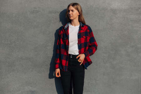 Portrait of a cheerful young woman dressed in casual clothes and a plaid shirt standing against a...
