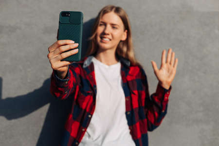 Happy young woman making face to face video calls with smartphone using online meeting app, social distancing, woman using mobile phone against gray street wall outdoors, remote work conceptの写真素材