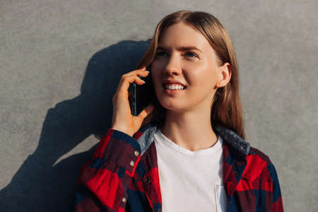 Beautiful smiling woman calling on a mobile phone on an open city street, young happy business woman in casual shirt talking against a gray city wall backgroundの写真素材