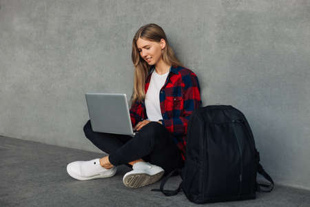 Happy young woman with backpack, in casual clothes, sitting on the floor with crossed legs and using...