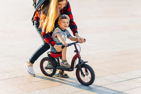 Little happy child learns to ride a bike with her mom in the park in the park, Happy young mom teaches her son to ride a bike in the park on a summer sunny dayの写真素材