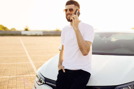 Handsome young business man talking on a mobile phone in the city near a white car, a man in glasses posing near a car in the city outdoorsの写真素材