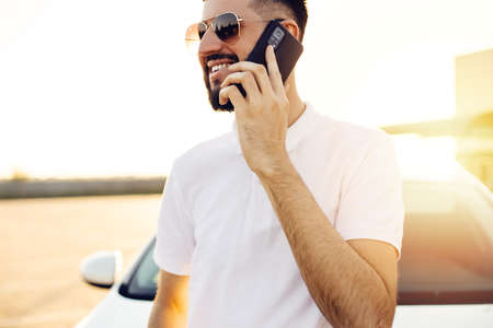 Handsome young business man talking on a mobile phone in the city near a white car, a man in glasses posing near a car in the city outdoorsの写真素材