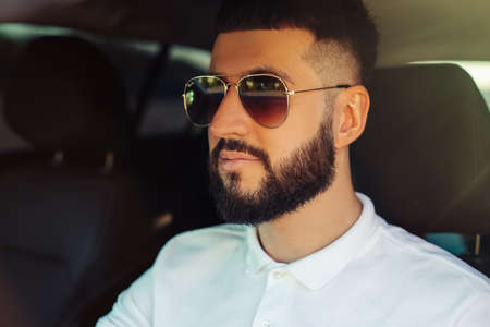 Handsome young man with beard wearing sunglasses smiles while driving a car, on a summer sunny dayの写真素材