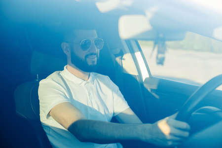 Front view, handsome young business man with beard sitting in car holding steering wheel smiling happily looking at roadの写真素材