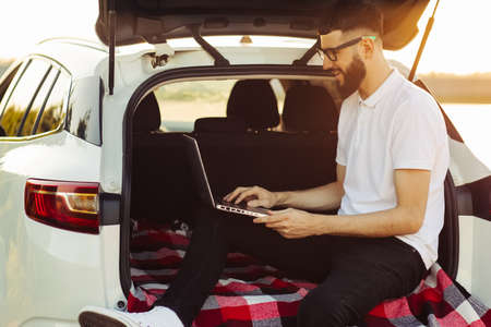Young man in sunglasses enjoying a trip in his car, sitting in the trunk of a car with a laptop, Travel concept, summer vacation, vacationの写真素材