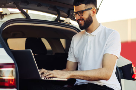 Young man in sunglasses enjoying a trip in his car, sitting in the trunk of a car with a laptop, Travel concept, summer vacation, vacationの写真素材