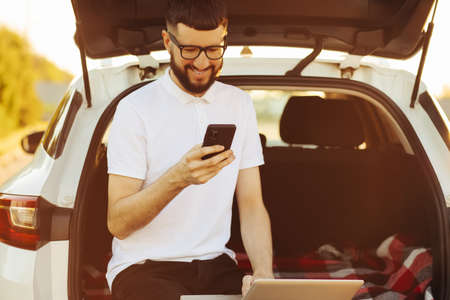 Young business man with laptop uses mobile phone while sitting in car trunk on summer vacation, travel concept, remote workの写真素材