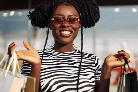 Portrait of a carefree fashionable African American woman wearing glasses, with shopping bags, happy...