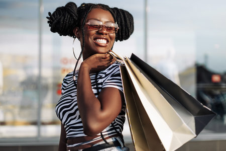 Portrait of a carefree fashionable African American woman wearing glasses, with shopping bags, happy woman with bags coming out of the store, shopping, Black Friday, salesの写真素材