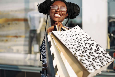 Young smiling african american woman, wearing sunglasses, with shopping bags in mall, shopping, black friday, discounts, sale conceptの写真素材