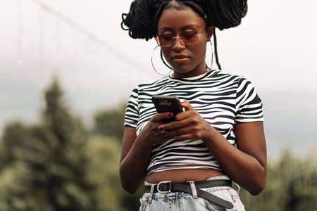 Close up portrait of beautiful young african american woman with afro hair wearing sunglasses walking outdoors using mobile phoneの写真素材