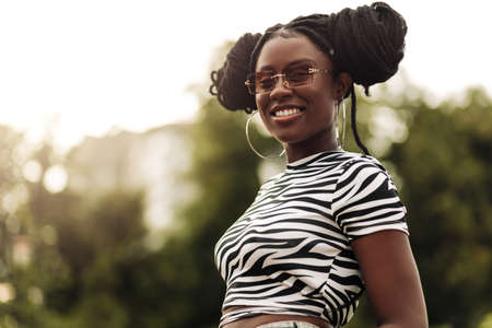 Portrait of young beautiful happy african american woman with hairstyle and sunglasses walking in city, smiling black woman outdoors in parkの写真素材