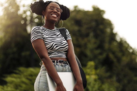 Beautiful young African American female student, with a backpack and laptop outdoors, College student with a lot of booksの写真素材