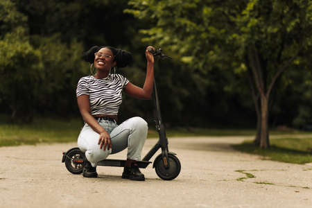 Beautiful African woman wearing sunglasses sits on her electric scooter in the park outdoorsの写真素材
