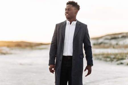 Handsome stylish black man in classic white shirt, trousers and coat, posing outdoors at sunset,...