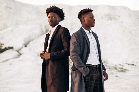 Two handsome stylish African American men, in classic suits and cardigans, two young men posing in fashionable clothes outdoors against the background of a beautiful landscape of mountains at sunsetの写真素材