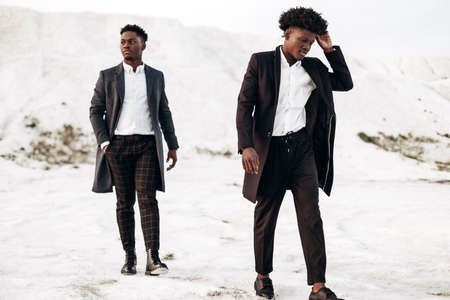 Two handsome African black men, walking and posing in classic clothes, down the street in fashionable coats, at sunset against the backdrop of white chalk mountainsの写真素材