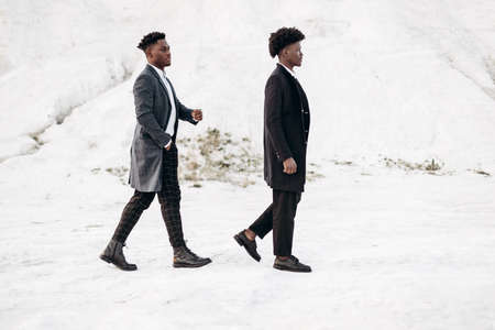 Two handsome African black men, walking and posing in classic clothes, down the street in fashionable coats, at sunset against the backdrop of white chalk mountainsの写真素材