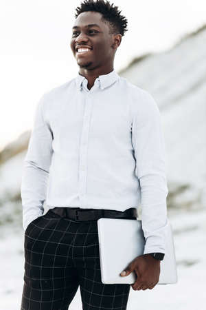 Business fashionable handsome african american man in white shirt posing outdoors holding a laptop,...