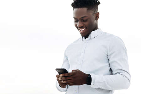 Happy fashionable curly African man, in white shirt, using mobile phone, outdoors on the street,...