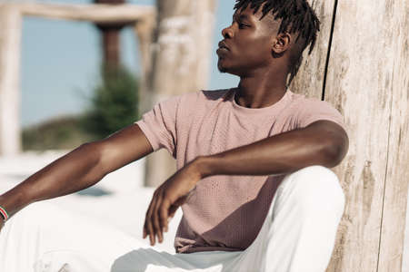 Fashionable african american man, in stylish t-shirt and white pants, sits cross-legged in the sandy...