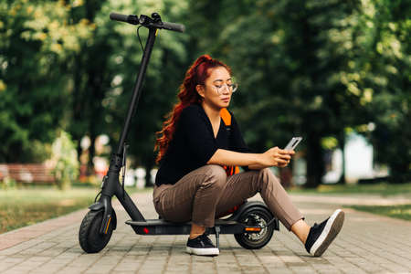 Happy attractive asian female, wearing glasses standing with electric scooter outdoors in park, woman with red backpack using mobile phoneの写真素材