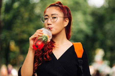 Portrait of smiling asian woman drinking cocktail while walking along city street, woman walking and enjoying warm day in parkの写真素材