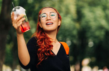 Smiling beautiful Asian woman, holding delicious fruit cooling cocktail while walking down the street, Beautiful young woman enjoying summer vacationの写真素材