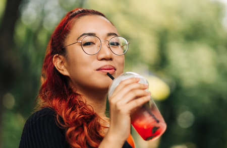 Smiling beautiful Asian woman, holding delicious fruit cooling cocktail while walking down the street, Beautiful young woman enjoying summer vacationの写真素材