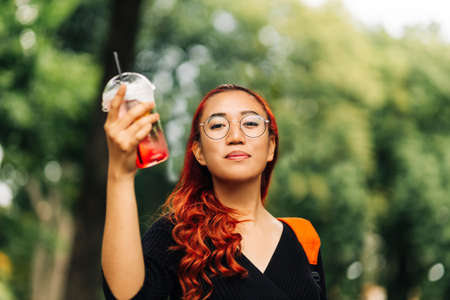 Smiling beautiful Asian woman, holding delicious fruit cooling cocktail while walking down the street, Beautiful young woman enjoying summer vacationの写真素材