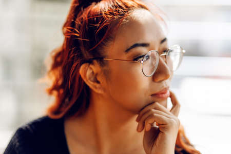 pensive Asian woman, with glasses looks out the window, with a hand on her chin, thinking about a question, obsessive expression. Smiling with thoughtful faceの写真素材