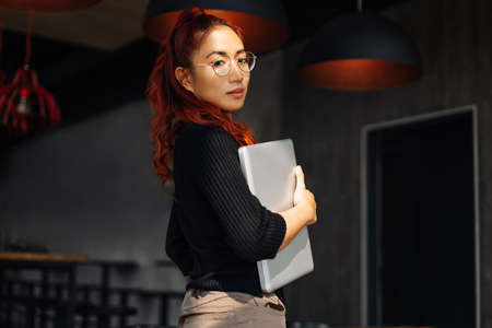 Portrait of young Korean Asian female student wearing glasses, with laptop, Young woman standing in...