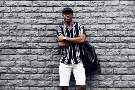 handsome african man in a cap and shirt with a backpack, against the background of a brick black wallの写真素材