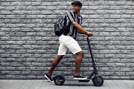 African american man with scooter on black brick wall background, guy with backpack, young student with electric scooterの写真素材