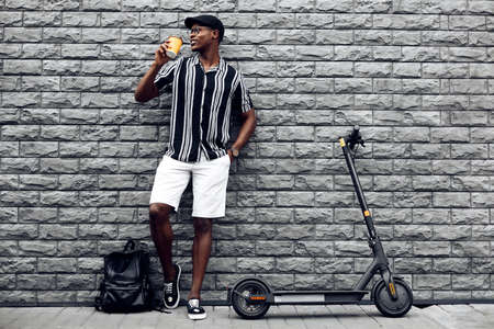 stylish african american man with paper cup of coffee or tea in hand, and electric scooter, stands against black wall background, outdoor recreationの写真素材