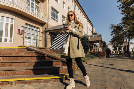 beautiful women with shopping bags in a beige raincoat and a phone in their hands makes online...