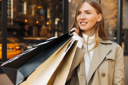 beautiful woman in a beige raincoat and shopping bags in front of a glass building, and lights. autumn and shopping, sale.の写真素材