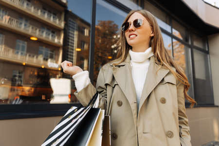 a beautiful woman in the sun of goggles in a beige raincoat and shopping bags in her hands, stands in front of a glass building, and lights. autumn theme: shopping.の写真素材