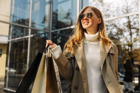 blonde european woman in sunglasses in a beige raincoat and with shopping bags in her hands stands in front of a glass building, a shopping center, shopping in the city, outdoorsの写真素材