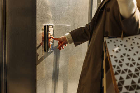 close-up of a finger presses the elevator button. a woman with shopping bags in a beige raincoat...