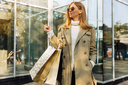 beautiful blonde in sun goggles and shopping bags stands on against a glass building, in a beige raincoat. concept: autumn shopping, black friday, novemberの写真素材