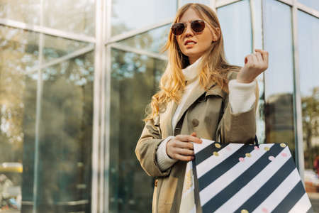 a beautiful blonde in sun goggles and shopping bags stands on against a glass building, in a beige...