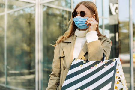 a woman holds a protective mask from coronavirus with her hand, and shopping bags in her hands, in a...