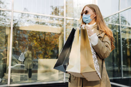 stylish woman in a protective mask against coronavirus, and shopping bags in her hands, in a beige...