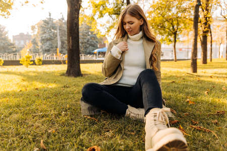 beautiful woman in a beige raincoat sits on the grass in the park, against the background of autumn...