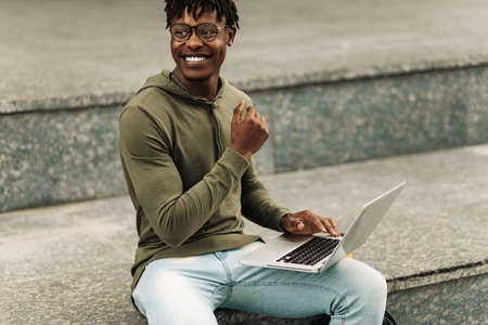 Happy Black African man, with glasses working on laptop outdoors, curly haired man sitting on stairs outdoors working on laptopの写真素材