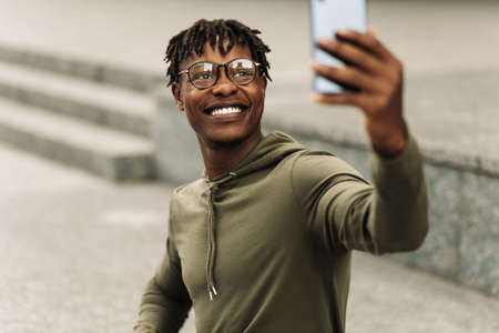 Excited happy black african man, wearing glasses, taking selfie on camera, wearing casual fashion clothes on a walk in the city outdoorsの写真素材