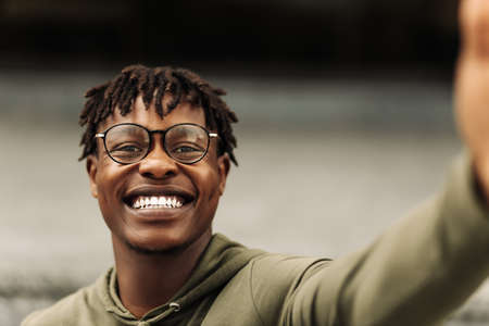 Excited happy black african man, wearing glasses, taking selfie on camera, wearing casual fashion...